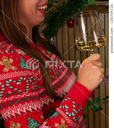 shot of young woman smiling while holding a glass with white wine or champagne wearing a red Christmas sweater 120704316