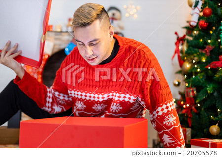 Christmas day concept. handsome man sitting surprised and opening gift box with christmas tree background. Festive atmosphere party. 120705578