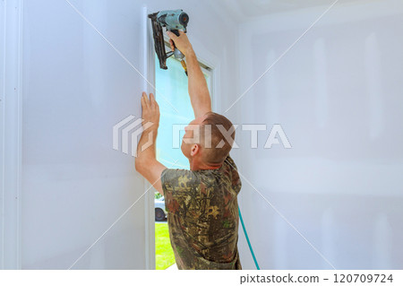 Construction worker is attaching trim to wall with nail gun in around window 120709724