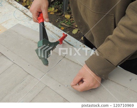 A man uses a riveter to fasten together gray thin metal plates that lay on a rusty blue table 120710969