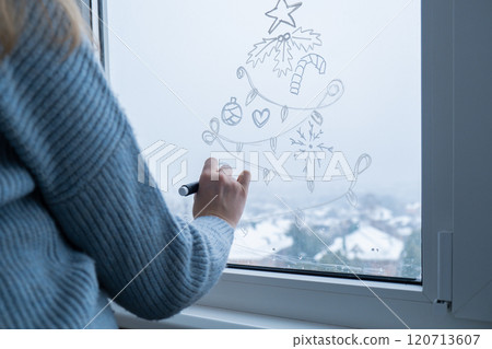 Creative enthusiastic unrecognizable woman in sweater paint window with white marker. Christmas tree decoration on big clear window. Snowy winter outside 120713607