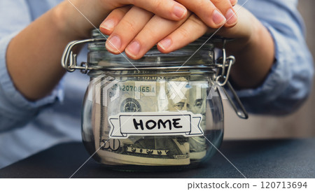 Female hands holding Glass jar full of American currency dollars cash banknote with text HOME. Preparation saving money. Moderate consumption and economy Female hands holding Glass jar full of American currency dollars cash banknote with text HOME. Preparation saving money. Moderate consumption and economy 120713694