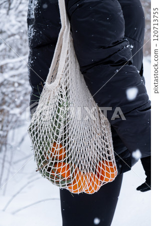 Unrecognizable woman holding Mesh shopping bag with tangerines in snowy winter forest outdoor. Go green concept eco-friendly sustainable Christmas. Copy space for text. Responsible Unrecognizable woman holding Mesh shopping bag with tangerines in snowy winter forest outdoor. Go green concept eco-friendly sustainable Christmas. Copy space for text. Responsible 120713755