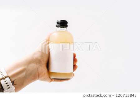 Woman hand holding small transparent glass bottle with black cap and white label, filled kombucha of yellow Woman hand holding small transparent glass bottle with black cap and white label, filled kombucha of yellow 120713845