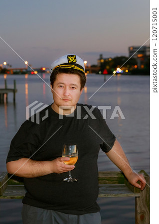 Man stands by water at dusk, wearing captain hat holding glass of drink sun sets, casting warm glow over scene reflecting on water. 120715001