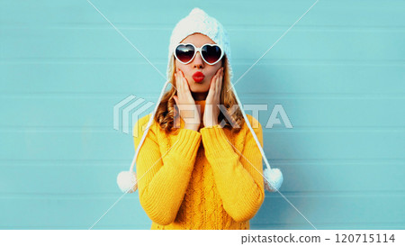 Winter portrait young woman blowing kiss wearing yellow knitted sweater and white hat on blue wall Winter portrait young woman blowing kiss wearing yellow knitted sweater and white hat on blue wall 120715114