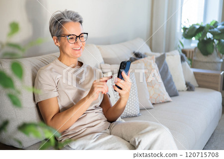 Mature beautiful middle-aged woman with gray hair and poor eyesight sits on the couch Mature beautiful middle-aged woman with gray hair and poor eyesight sits on the couch 120715708