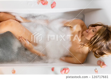 A young beautiful girl drinks wine while lying in a bath with foam and petals.  120717681