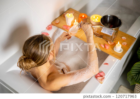 A young beautiful girl drinks wine while lying in a bath with foam and petals. Candles and plants 120717963