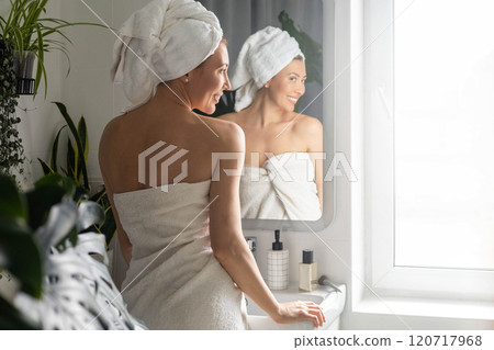 A young beautiful woman in a white terry towel admires herself in front of a mirror in her bathroom. A young beautiful woman in a white terry towel admires herself in front of a mirror in her bathroom. 120717968