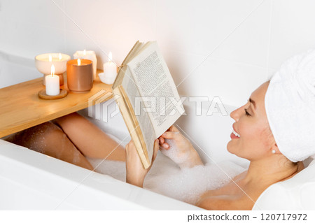 A young beautiful woman lies in a bath with white foam. Reading a book and enjoying a bath. A young beautiful woman lies in a bath with white foam. Reading a book and enjoying a bath. 120717972