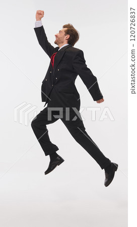 Young businessman in black suit and red tie leaps joyfully in mid-air, showcasing energy and success. against white studio background. 120726837