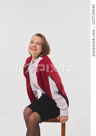Happy young businesswoman in formal attire, wrapped in red sweater, sits on wooden stool, looking upward with smile against white studio background. Happy young businesswoman in formal attire, wrapped in red sweater, sits on wooden stool, looking upward with smile against white studio background. 120726840