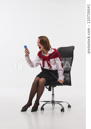 Young professional, dressed in white and black attire with red sweater, shouts at her smartphone against white studio background. Young professional, dressed in white and black attire with red sweater, shouts at her smartphone against white studio background. 120726841