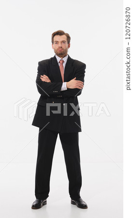 Stylish young man in black suit with red tie, standing confidently with arms crossed against white studio background. 120726870