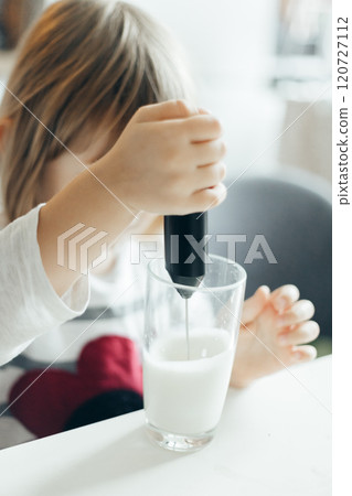 Pre-school child makes frothy milk with a hand mixer in a glass cup in the kitchen 120727112