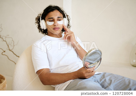 Man dressed in casual clothes with headphones on head sitting in comfortable chair holding mirror and adjusting eye patch Man dressed in casual clothes with headphones on head sitting in comfortable chair holding mirror and adjusting eye patch 120728004