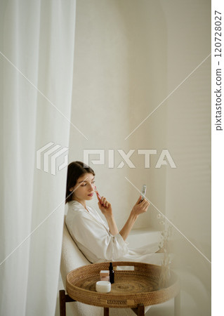 Vertical shot of young adult woman massaging face with face roller while looking in mirror 120728027