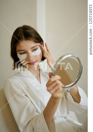 Vertical shot of young adult Caucasian woman admiring herself in mirror while adjusting collagen eye patches to decrease pussiness 120728055