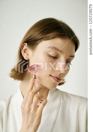Vertical shot of Caucasian woman using rose pink face roller to massage face Vertical shot of Caucasian woman using rose pink face roller to massage face 120728079