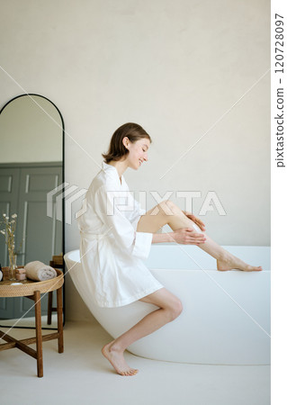 Vertical shot of smiling Caucasian woman in white silk bathrobe smearing perfumed cream while sitting in aesthetic bathroom at home Vertical shot of smiling Caucasian woman in white silk bathrobe smearing perfumed cream while sitting in aesthetic bathroom at home 120728097