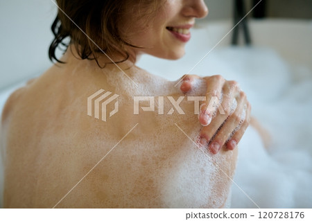 Medium close up of unrecognizable smiling woman washing her body with aromatic shower gel while sitting in hot bathtub 120728176