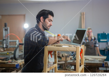 young carpenter caucasian man with laptop make drawing on wood to design at workspace. craftsman profession in wood factory young carpenter caucasian man with laptop make drawing on wood to design at workspace. craftsman profession in wood factory 120728264
