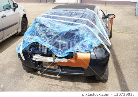 An accident-damaged car stands wrapped in film in a parking lot. Broken rear doors and bumper are visible. An accident-damaged car stands wrapped in film in a parking lot. Broken rear doors and bumper are visible. 120729033
