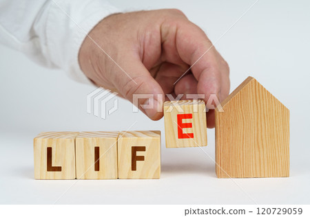On a white surface there is a model of a house, next to it a man has placed cubes with the inscription - life On a white surface there is a model of a house, next to it a man has placed cubes with the inscription - life 120729059