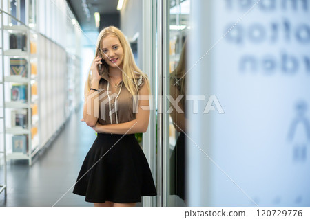 Attractive girl talking on phone in library. Female converse with mobile phone. Beautiful caucasian woman with blond hair talking telephone. Students education conversation. 120729776