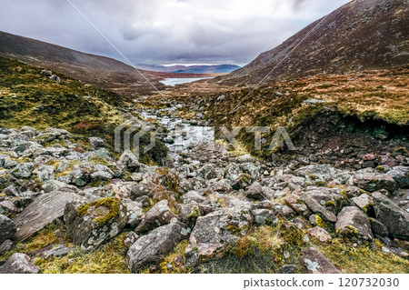 A stream flowing through a rocky valley. The stream is surrounded by rocks, covered in moss and grass. The valley is narrow and steep, with hills rising up on either side. The sky is overcast. 120732030