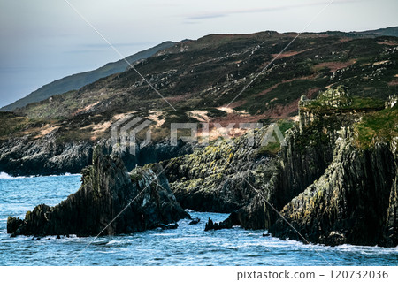 Rugged cliffs rise majestically above the calm blue waters, contrasting with the lush green hills of cloudy Ireland. Rugged cliffs rise majestically above the calm blue waters, contrasting with the lush green hills of cloudy Ireland. 120732036