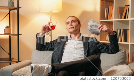 Rich man. Happy relaxation. Successful handsome young guy in modern suit holding money and glass of red wine sitting in office on lighted yellow background. Rich man. Happy relaxation. Successful handsome young guy in modern suit holding money and glass of red wine sitting in office on lighted yellow background. 120733063