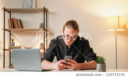 Career man. Technology help. Successful young handsome guy in glasses in black shirt holding smartphone before laptop sitting in office online using gadget. 120733141