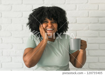 African American joyful woman laughing while holding a gray mug, expressing happiness and positivity. Carefree moments, laughter, and beauty of authentic emotions 120735405