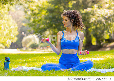 Sportive woman exercising weightlifting with dumbbells sitting on yoga mat in park on sunny day Sportive woman exercising weightlifting with dumbbells sitting on yoga mat in park on sunny day 120736803
