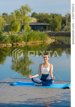 Caucasian young woman sitting on mat in lotus position, relaxing near pond in park on summer day 120736815