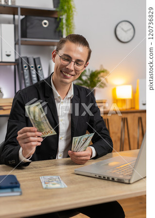 Happy young Caucasian businessman counting money cash and using laptop computer in home office Happy young Caucasian businessman counting money cash and using laptop computer in home office 120736829