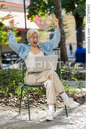 Overjoyed happy young woman working on laptop screaming in delight raises hands in city street 120736942