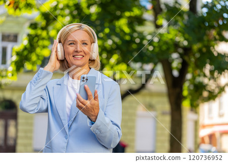 Happy carefree mature businesswoman with smartphone listening to music in headphones on city street Happy carefree mature businesswoman with smartphone listening to music in headphones on city street 120736952