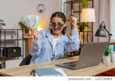 Young woman freelancer putting on party glasses holding fan of cash sitting at home office table Young woman freelancer putting on party glasses holding fan of cash sitting at home office table 120736963