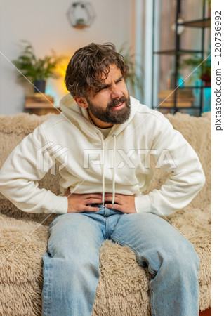 Young Indian man sitting on sofa feeling sudden strong abdominal stomach ache, gastritis problem 120736992