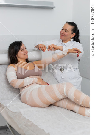 Cosmetologist warping woman hand with compression elastic bandage body mask regeneration weight loss 120737043