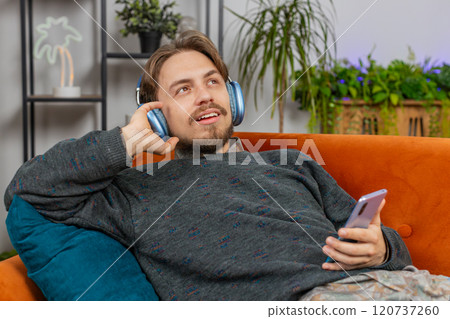 Happy man in wireless headphones relaxing listening favorite rock n roll music lying on home couch Happy man in wireless headphones relaxing listening favorite rock n roll music lying on home couch 120737260