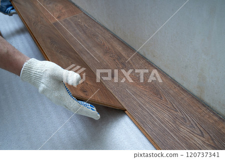 Using gloves, the person is meticulously installing a hardwood floor with planks, applying wood stain, varnish, and building material like plywood 120737341