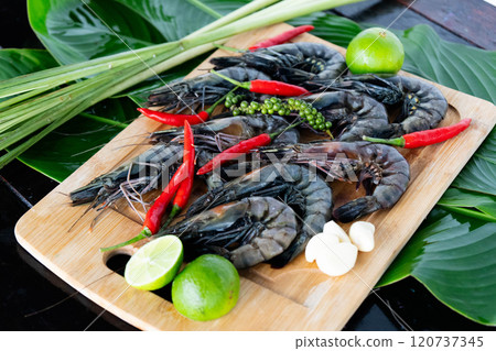 Brightly colored tiger shrimp rest on banana leaves alongside fresh herbs, chili peppers, and lime, showcasing the essence of Asian culinary traditions and vibrant flavors. 120737345