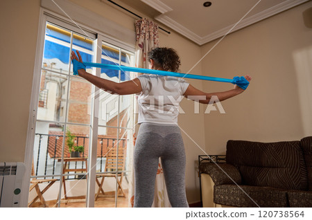 Woman exercising with resistance band at home by large window 120738564