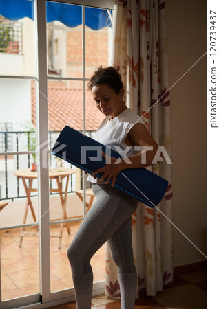 Woman holding yoga mat near a bright home window indoors Woman holding yoga mat near a bright home window indoors 120739437