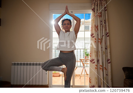 Woman practicing yoga tree pose in a bright living room Woman practicing yoga tree pose in a bright living room 120739476
