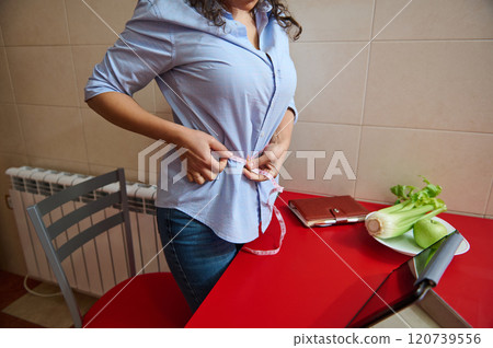 Woman measuring waist in kitchen with healthy food on table 120739556
